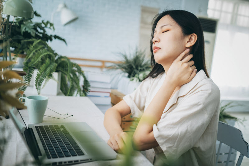 Woman With Tech Neck Symptoms Sitting at Table in Front of Laptop Holding Neck in Pain