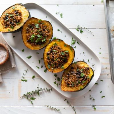 Einkorn Wheat Berries Stuffed Squash on Long, White Serving Plate on top of a White Wood Table Top Scattered With Fresh Herbs