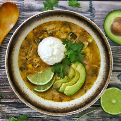 An Overhead View Shows a Bowl of Green Chicken Chili Topped With Avocado Slices, Lime Wedges, Cilantro and Sour Cream.