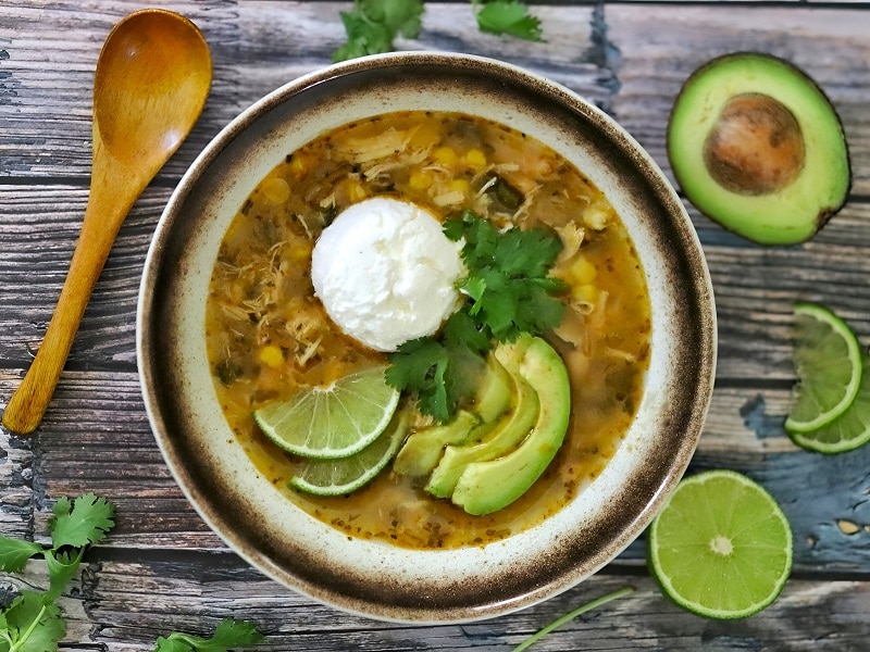 An Overhead View Shows a Bowl of Green Chicken Chili Topped With Avocado Slices, Lime Wedges, Cilantro and Sour Cream.