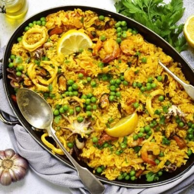 Classic Spanish Valencian paella from rice with seafood and green peas in a paella pan on a light stone background scattered with lemons and garlic