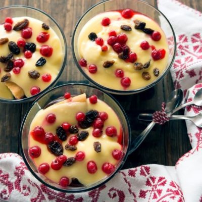 Three glass bowls of homemade vanilla pudding sprinkled with small berries sitting atop a textural wooden table next to a napkin with raised embroidery