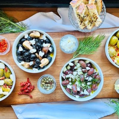 Marinated olives four different ways in four bowls set out on a wooden charcuterie board with toasted bread