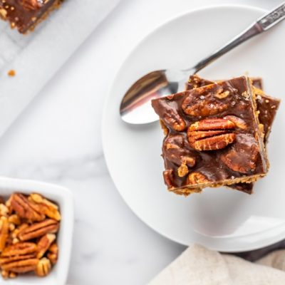 Salted Caramel Pecan Pie Bars are Stacked High on a White Plate With Whole Pecans in a Small Dish Off to the Side.
