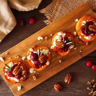 Round slices of roasted sweet potatoes sprinkled with goat cheese, pecans, cranberries and thyme on a rustic wooden serving board, pinecones and linen surround it