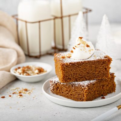 Two Slices of Gingerbread Cake Rest On a Small Plate Topped With Powdered Sugar and a Dollop of Whipped Cream.