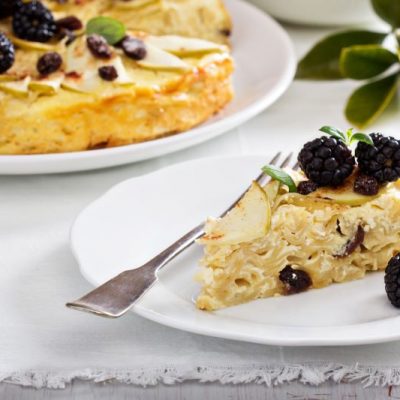 High-Protein Gluten-Free Noodle Kugel: One Slice on a Plate with Blackberries, Apple Slices & Mint Leaves. A Large Round Kugel on Serving Platter is in the Background