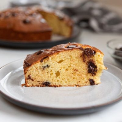 A Slice of Chocolate Chip Ricotta Cake on a Stoneware Plate Set in Front of an 8-inch Cake Dotted with Chocolate Chips