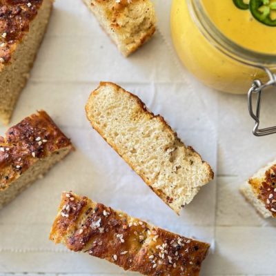 Slices of Pretzel Focaccia Sprinkled With Everything Seasoning & Flake Salt Placed Around a Jar of Vegan Cheese Sauce