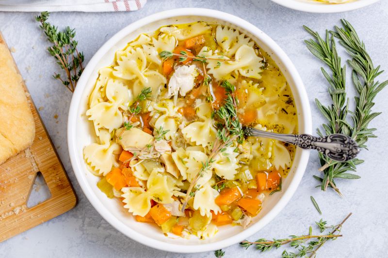 Comforting Homemade Chicken Noodle Soup in a Large Bowl With a Waiting Spoon, Sprigs of Rosemary & Thyme Surround it