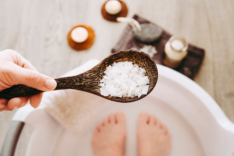 Magnesium Flakes Benefits: Soothe Muscles, Relax Mind & Body A Woman Holds a Scoopful of Magnesium Flakes Over a Foot Bath, Representing Magnesium Flakes Benefits.