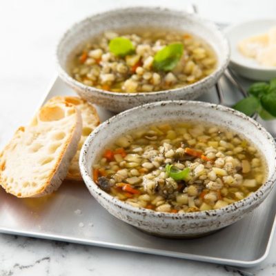 Two Earthenware Bowls of Mushroom Barley Soup Topped with Cilantro Leaves on Silver Tray with Slices of Crusty Bread