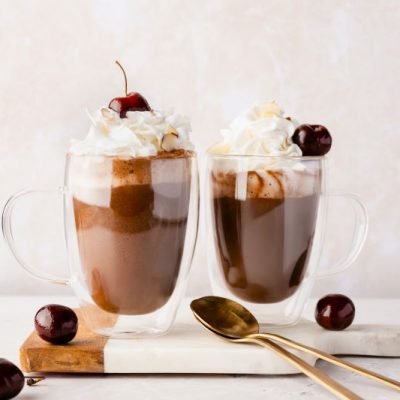 Two Tall Glass Mugs of Dairy-Free Cherry Almond Hot Chocolate with Whopped Cream and a Cherry on Top with two Gold Long-Handled Spoons Nearby