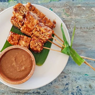Four Skewers of Chickpea Tempeh Satay Arranged on a Green Banana Leaf on a White Plate With a Bamboo Bowl of Sunflower Dipping Sauce