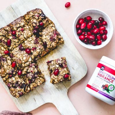 A Slab of Sliced Oatmeal Bars With Cranberries on Wooden Cutting Board with a Jar of Spectrum Vibrance Superfood Powder Nearby