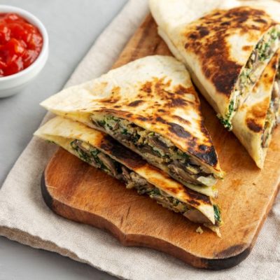Goat Cheese, Spinach & Mushroom Quesadilla Cut into Wedges on Wooden Serving Board with a Bowl of Salsa Nearby