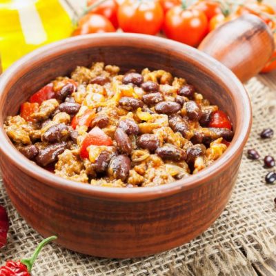 Vegan Chili with Tofu & Beans in Brown Earthenware Handled Crock on Burlap Placemat with Tomatoes & Chili Peppers Strewn Around