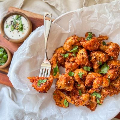 Maple-Sriracha Cauliflower Wings on Parchment Atop Wooden Appetizer Board with Bowls of Chopped Cilantro & Ranch Dressing for Dipping