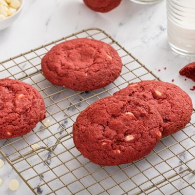 Red Velvet White Chocolate Chip Cookies Sit on a Cooling Rack Next To White Chocolate Chips and a Glass of Milk.