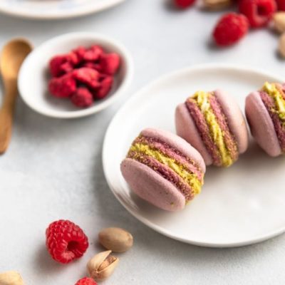 Three Raspberry Vegan Macarons on a Round Dessert Plate Surrounded by Raspberries and Pistachios