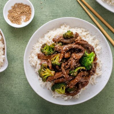 Homemade beef and broccoli stir fry over rice in large round serving bowl on green counter with chopsticks and a bowl of sesame seeds nearby
