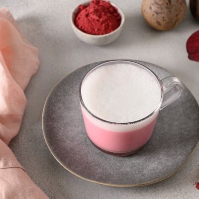 Pink Beetroot Latte in Glass Mug Set on Light Grey Saucer Next to a Pink Napkin. Beetroot Powder is in a Bowl Nearby next to several Fresh Beets