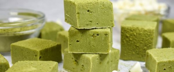 Close-up of White Chocolate Matcha Fudge Squares on Countertop Surrounded by White Chocolate Chips and a Glass Bowl of Matcha Powder