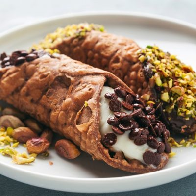Two Gluten Free Cannolis Sit on a Plate, One Topped With Pistachios and the Other Topped With Mini Chocolate Chips.