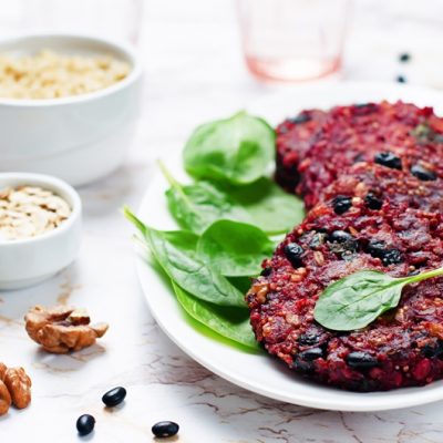 A White Platter With Four Black Bean and Quinoa Burgers Sits Beside Fresh Spinach, Rolled Oats and Walnuts.