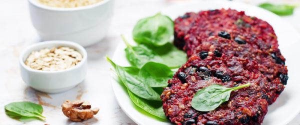 A White Platter With Four Black Bean and Quinoa Burgers Sits Beside Fresh Spinach, Rolled Oats and Walnuts.