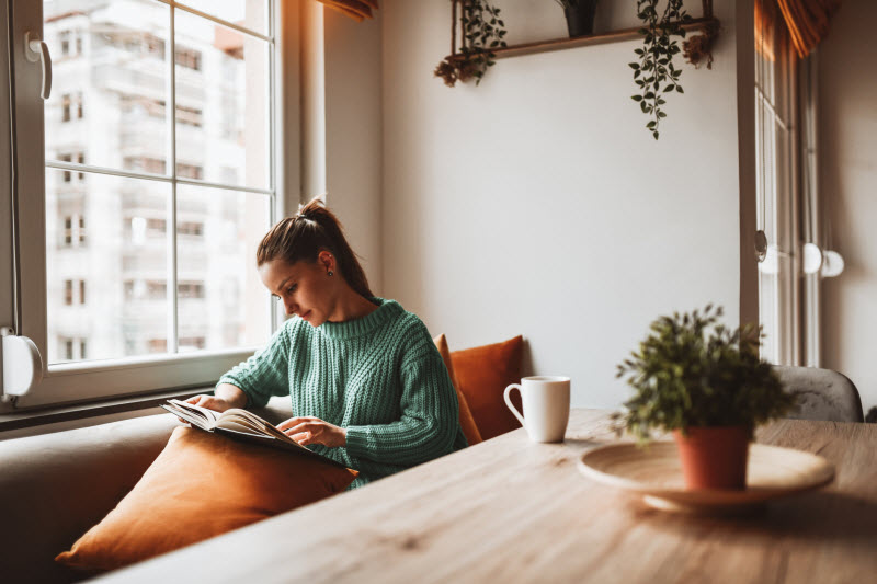 Woman Reading Book at Table by Window Risking Her Health With Indoor Living Lifestyle