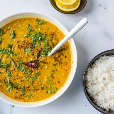 A Bowl of Red Lentil Dahl With Coconut Milk is Garnished With Cilantro, Pepper and Spices, Sits Beside a Bowl of White, Fluffy Basmati Rice.