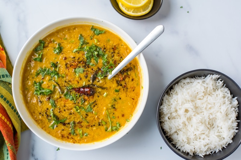 A Bowl of Red Lentil Dahl With Coconut Milk is Garnished With Cilantro, Pepper and Spices, Sits Beside a Bowl of White, Fluffy Basmati Rice.