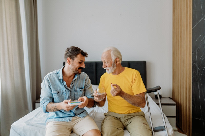 Concept of Supplements for Seniors Represented by Senior and Son Taking Vitamins While Sitting on Bed