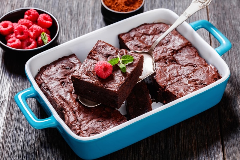 A Blue Baking Dish With Raspberry Brownies is Sliced into Squares With a Small Bowl of Fresh Raspberries on the Side for Garnish.