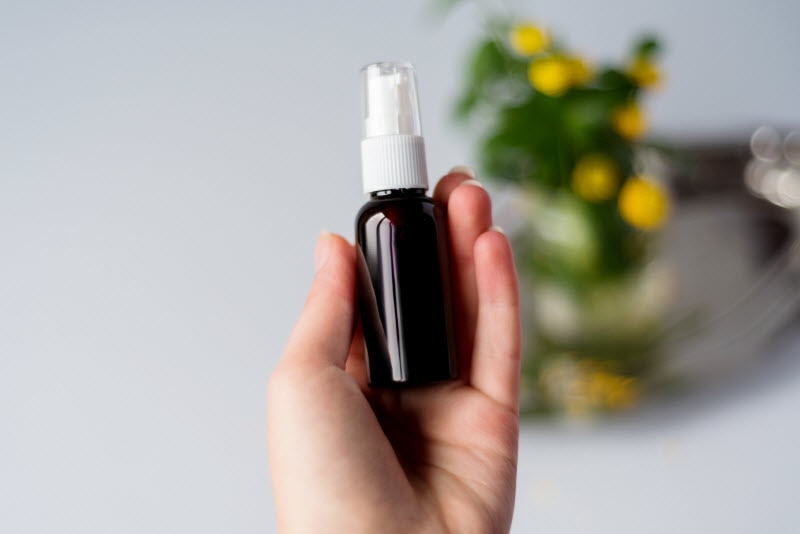 Close-up Shot of Hand Holding Dark Amber Vitamin Spray Bottle With Blurred Out Yellow Flowers in Background