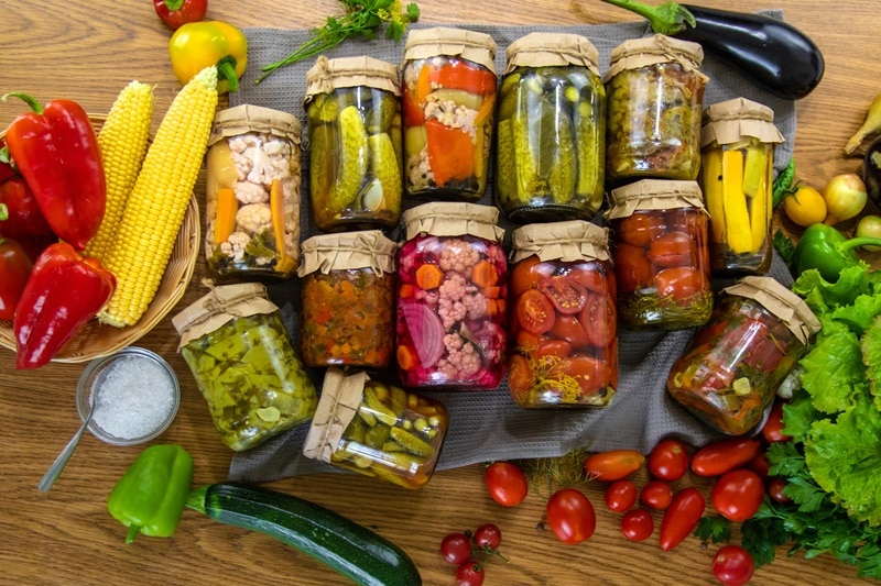 What Is Canning? Tips, Tools & Safety for Beginners A Collection of Preserved Vegetables is Laid Out on a Wooden Table Surrounded by Fresh Vegetables, Representing the Question, "What is Canning?"