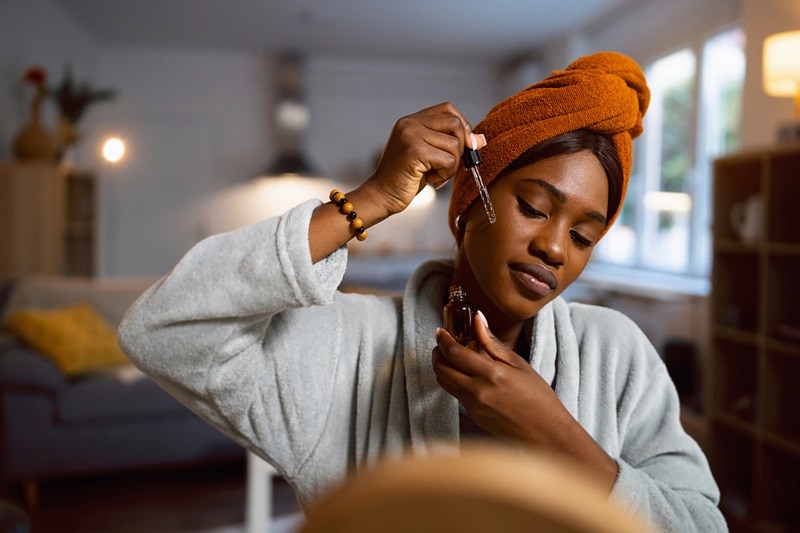 A Young Woman in a Robe Applies a Serum to Her Cheek, Representing Apricot Oil Benefits.