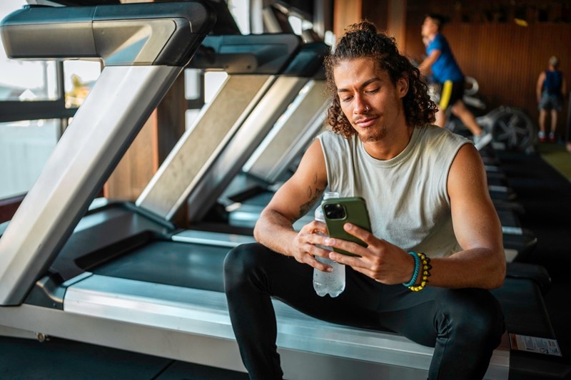 A Man Scrolls on His Phone During a Break at the Gym, Representing the Link Between Social Media and Eating Disorders.