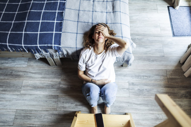 Overhead View of Exhausted Woman Sitting Near Bed With Hand on Forehead to Represent Concept of What is Adrenal Fatigue