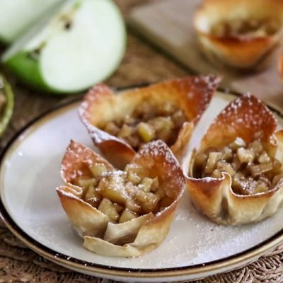 Powdered Sugar-Dusted, Browned Wonton Cups Filled with Cinnamon Apple Bits on a Dessert Plate Surrounded by Slices of Granny Smith Apples