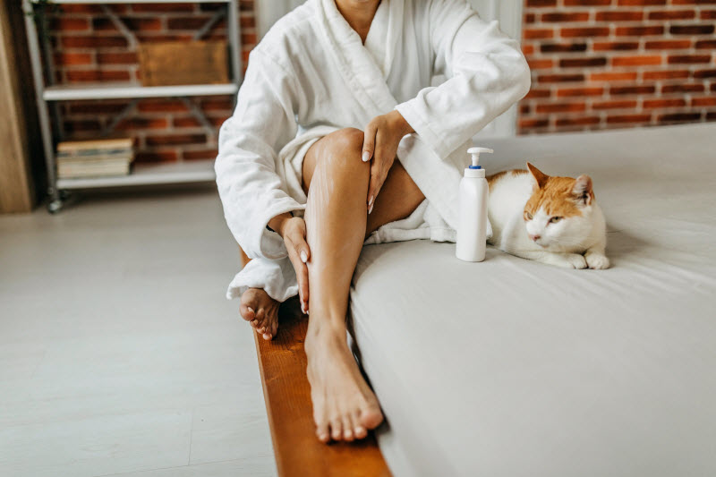 Woman Sitting on Floor Next to Cat Rubbing Lotion on Leg to Represent Concept of Types of Skin Rashes