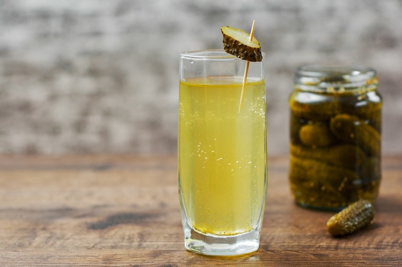 A Glass of Pickle Juice Sits Beside a Jar of Pickles, Representing the Question, "Is Pickle Juice Good for You?"