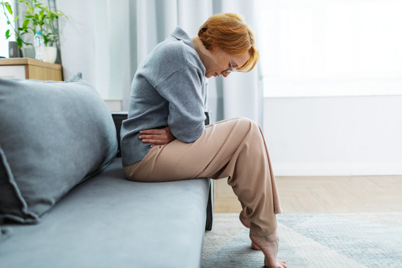 Woman Experiencing Menopause Bloating Holding Stomach in Pain While Sitting on Blue Sofa