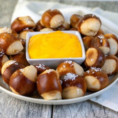 Salty Soft Pretzel Bites Surrounding Square Bowl of Creamy Vegan Cashew Cheese Dip on Weathered Wood Tabletop