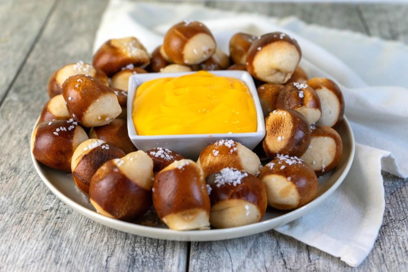 Salty Soft Pretzel Bites Surrounding Square Bowl of Creamy Vegan Cashew Cheese Dip on Weathered Wood Tabletop