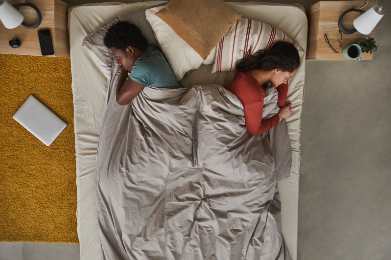 Couple Who Should Try the Scandinavian Sleep Method Sleeping Back to Back Under One Blanket in Small Bed