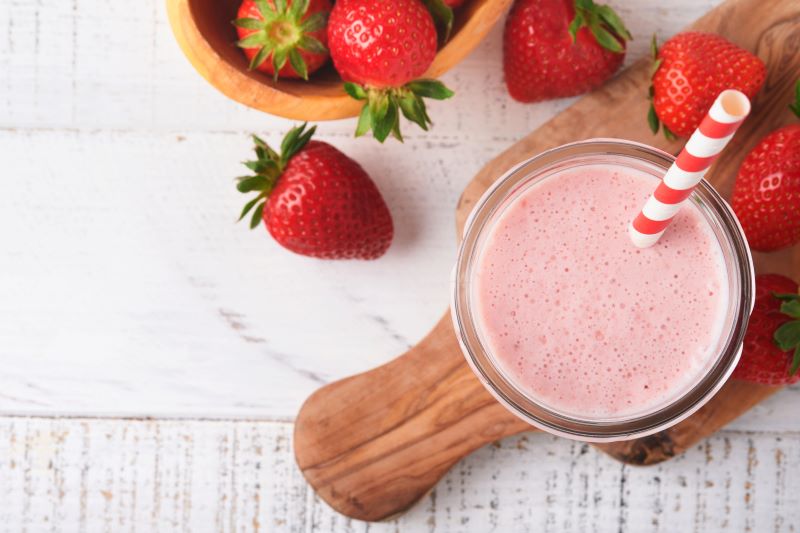 Top View of Glass of Frothy Strawberry Milk With a Straw in It, with Strawberries Dotting the Table