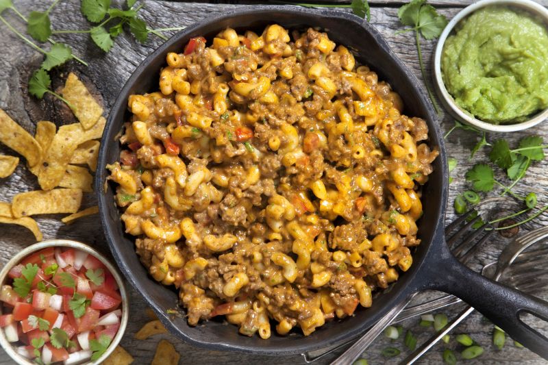Overhead Shot of Taco Macaroni and Cheese Loaded with Cheese-Covered Ground Beef in a Cast Iron Skillet Surrounded by Fresh Salsa & Guacamole