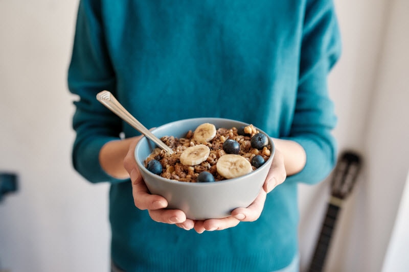 Person in Blue Sweater Holding Bowl of Cereal With Bananas and Blueberries to Represent Fibermaxxing Concept